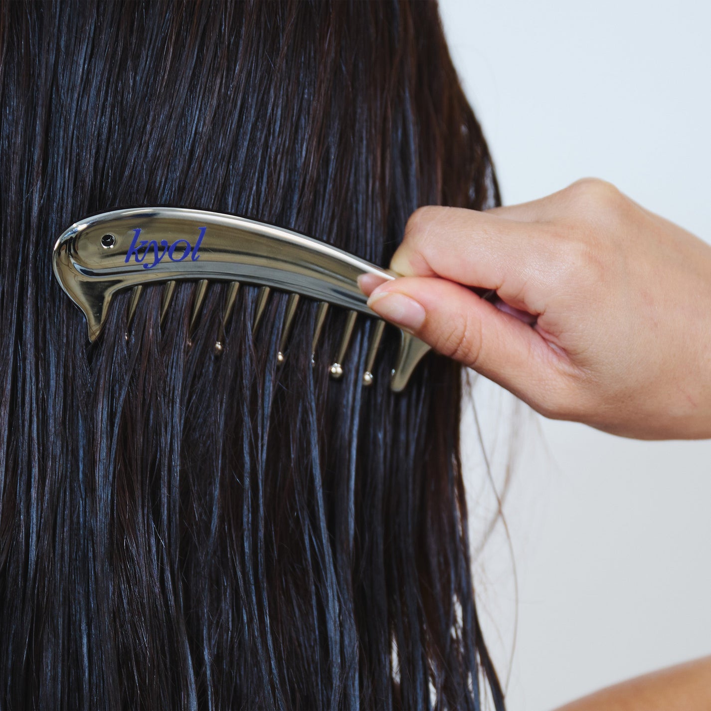 A close-up of a person's hand holding a shiny silver hair comb against wet, dark brown hair. The brand name 'Kyol' is printed in blue on the comb.