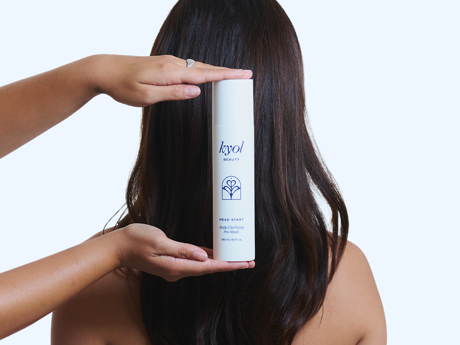 A woman with long dark brown hair stands with her back to the camera, holding a white bottle of Kyol Hair Care product in front of her head with both hands against a light blue studio background.